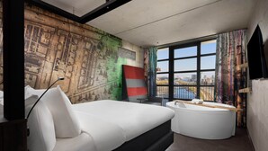 Une chambre d’hôtel moderne dotée d’un grand lit, d’un petit canapé, d’une baignoire et offrant une vue sur la ville grâce à de grandes fenêtres.