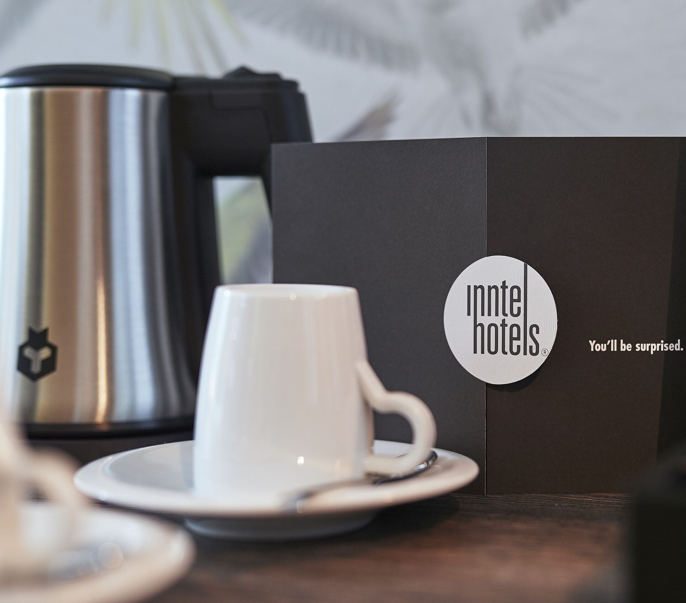 Eine weiße Tasse auf einem Untertasse, eine Edelstahl-Kaffeekanne und eine schwarze Box mit Logo und Schrift.