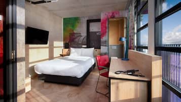 A modern hotel room with a bed, a desk, a chair, a TV, and large windows.