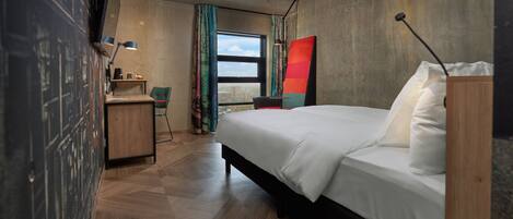 A modern hotel room with a large bed, a desk, a chair, and a view of the city through the window.