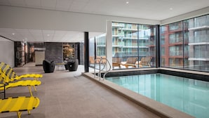 Une piscine intérieure moderne avec des parois en verre transparent, des sièges jaunes et noirs, et une vue sur un immeuble d’habitation.