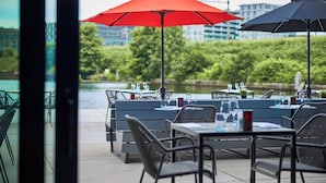 Un espace extérieur aménagé pour les repas, avec des tables et des chaises, des parasols rouges et noirs, et une vue sur une étendue d’eau et des bâtiments en arrière-plan.