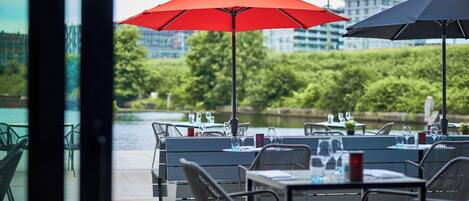 An outdoor dining area with tables and chairs, red and black umbrellas, and a view of a body of water and buildings in the background.