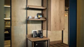 A compact kitchenette with a coffee maker, a small refrigerator, and a shelf with cups and a coffee pot.