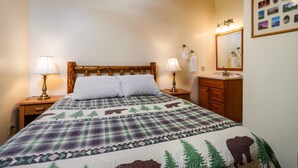 A bedroom with a bed, two bedside lamps, a wooden headboard, a nightstand, a dresser, a mirror, and a wall with framed pictures.
