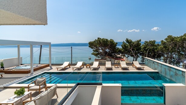 Een modern zwembad met ligbedden en een jacuzzi, met uitzicht op een heldere blauwe zee en verre bergen.