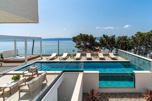A modern outdoor pool area with lounge chairs and a hot tub, overlooking a clear blue sea and distant mountains.