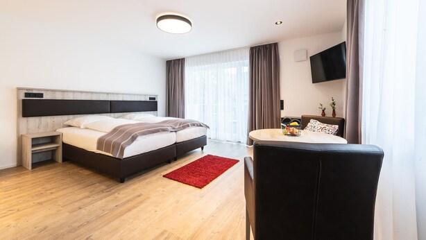A modern hotel room with a bed, a small table, a chair, a TV mounted on the wall, and a window with curtains. - Prim Loge (Balgheim)