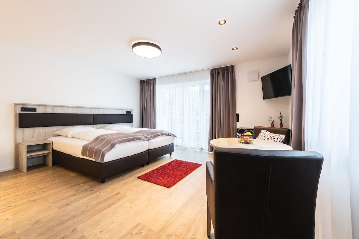 A modern hotel room with a bed, a small table, a chair, a TV mounted on the wall, and a window with curtains.