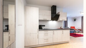 A modern kitchen with light wood cabinets, a black range hood, and a red rug in the living area. - Prim Loge (Balgheim)