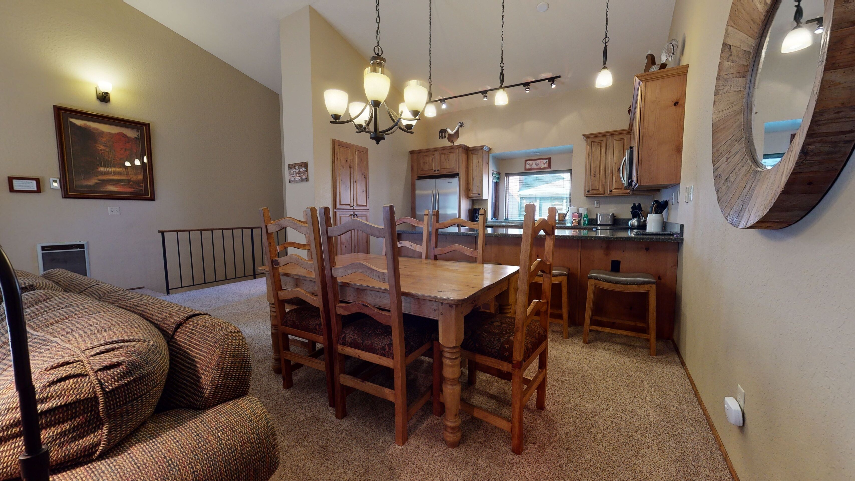 A dining area with a wooden table and chairs, a chandelier, and a kitchen in the background.