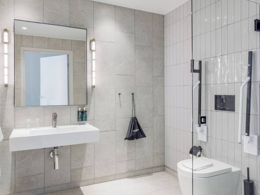 Ein modernes Badezimmer mit Waschbecken, Spiegel und Toilette in Weiß. Zwei Handtuchhalter und eine Duschkabine aus Glas sind ebenfalls vorhanden.