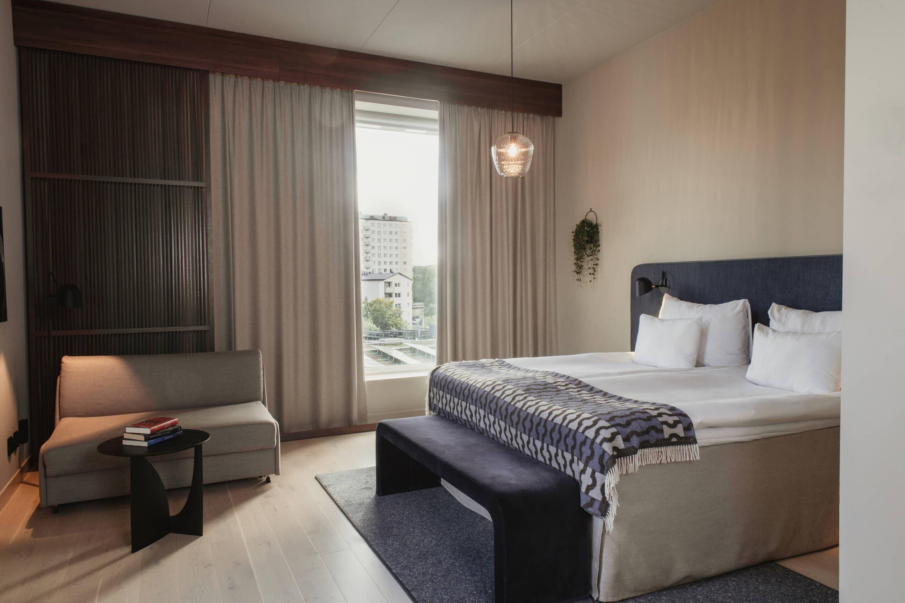 Ein modernes Hotelzimmer mit einem großen Bett, einer Couch, einem kleinen Tisch und Blick auf die Stadt durch das Fenster.