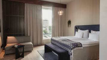 Ein modernes Hotelzimmer mit einem großen Bett, einer Couch, einem kleinen Tisch und Blick auf die Stadt durch das Fenster.
