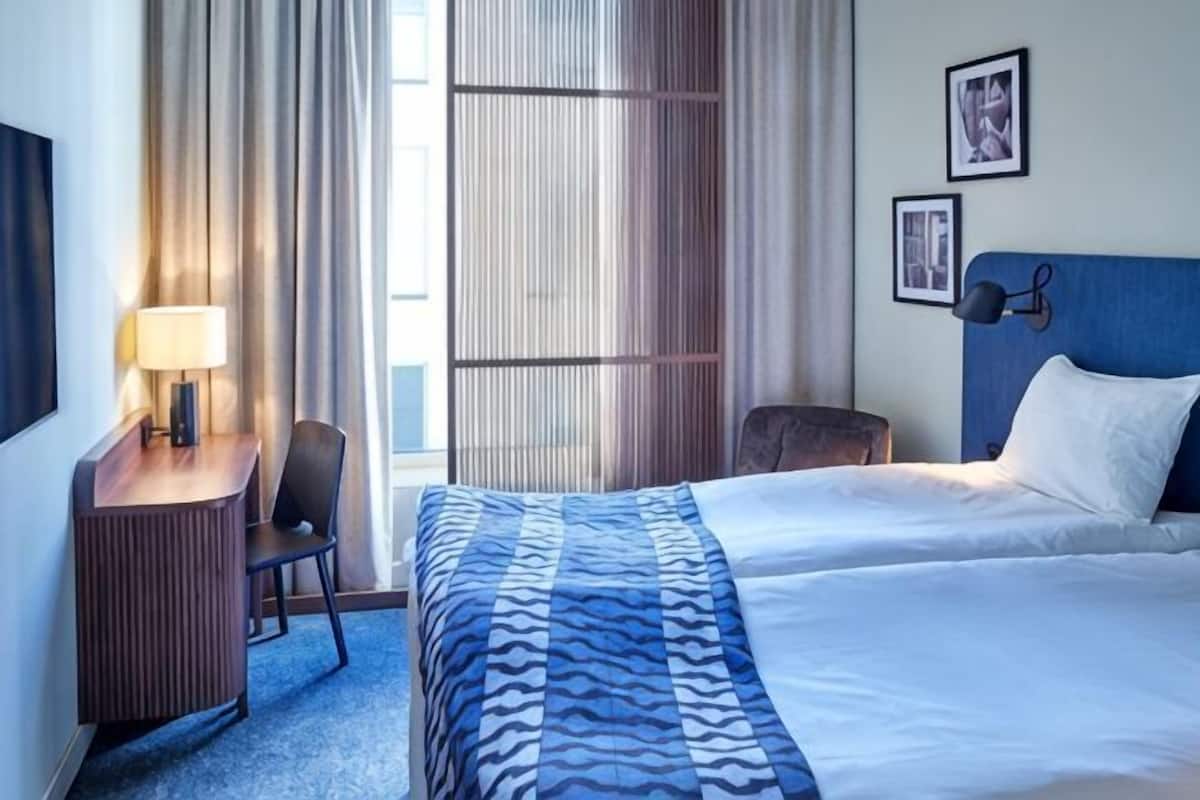 Una habitación de hotel con cama, escritorio, silla, lámpara y ventana con cortinas.