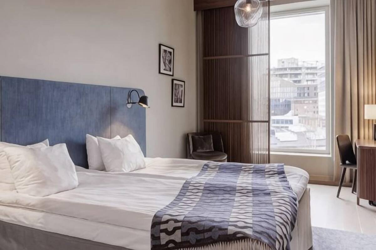 Habitación de hotel con una cama grande, un escritorio, una silla, una ventana que ofrece vistas a edificios y una lámpara fijada a la pared.