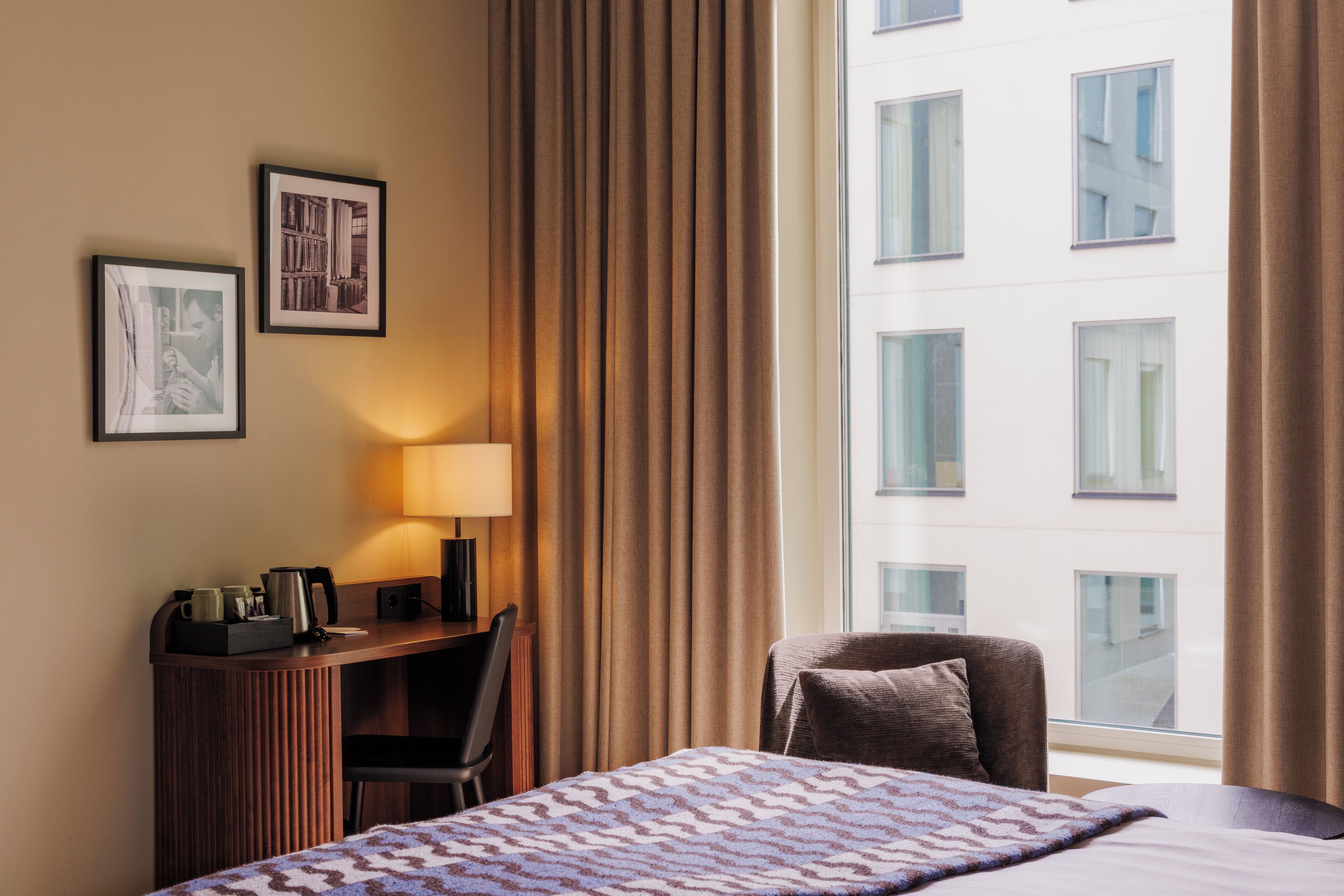 A hotel room with a bed, a desk with a lamp, a chair, and a large window with curtains.