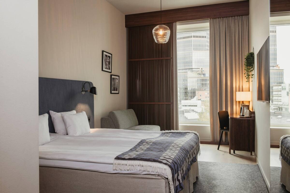 Habitación de hotel con cama, escritorio, silla, lámpara colgante y vista a la ciudad.