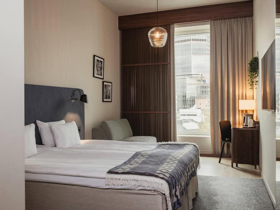 A hotel room with a bed, a desk, a chair, a hanging light fixture, and a view of the city.