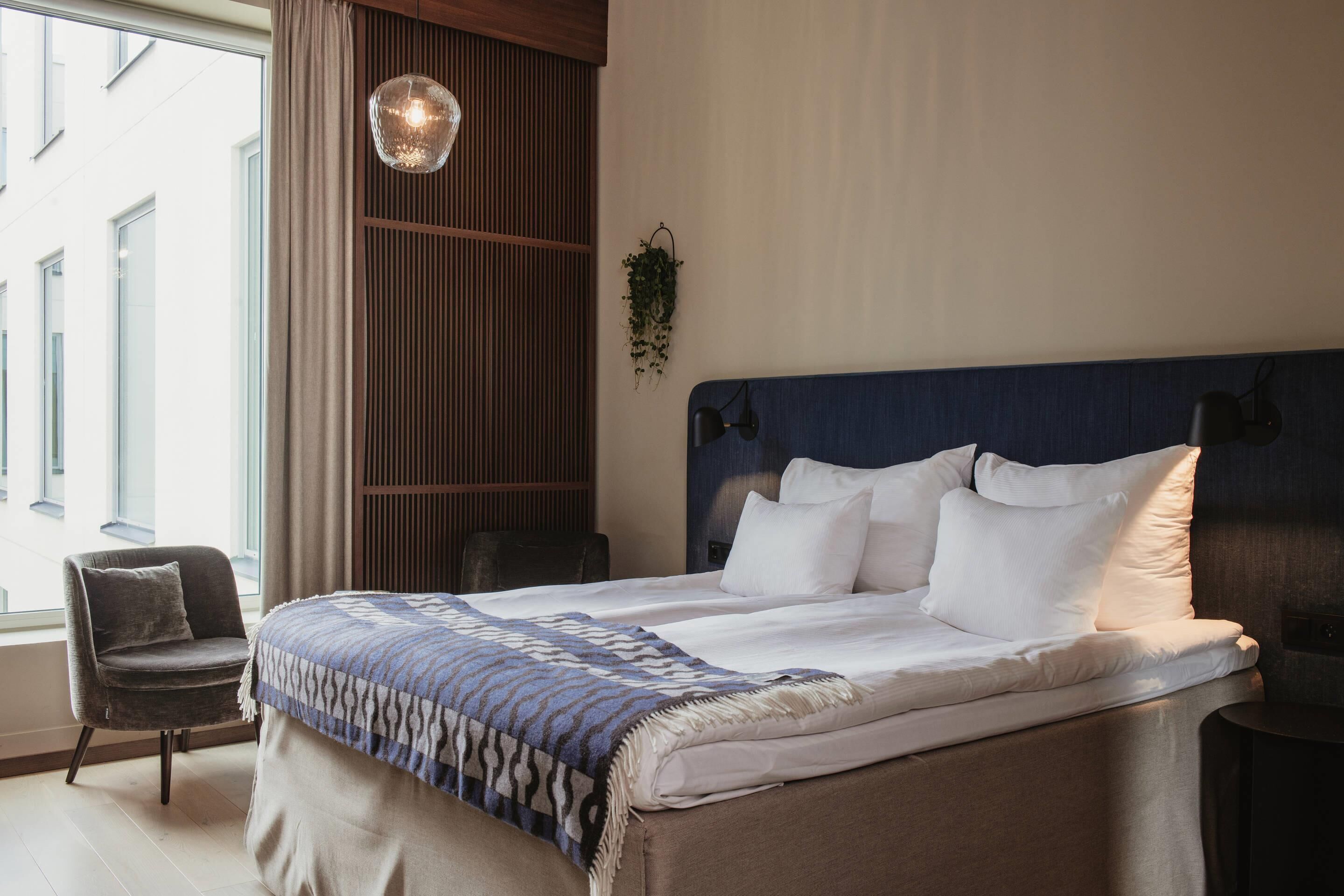 A hotel room with a large bed, a chair, a hanging plant, and a view of the outside.
