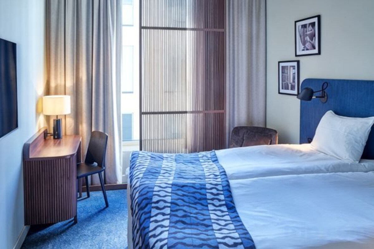 Una habitación de hotel con cama, escritorio, silla y ventana con cortinas.