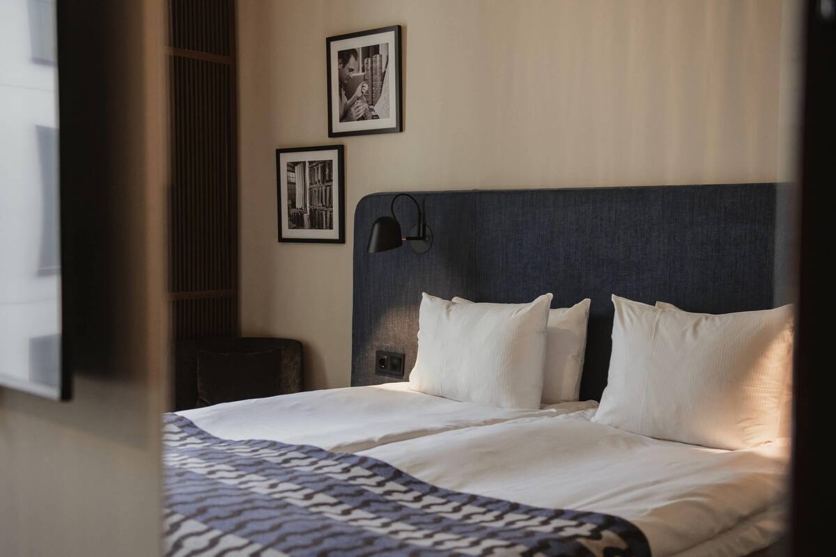 Habitación de hotel con cama, cabecera, mesita de noche y dos cuadros en la pared.