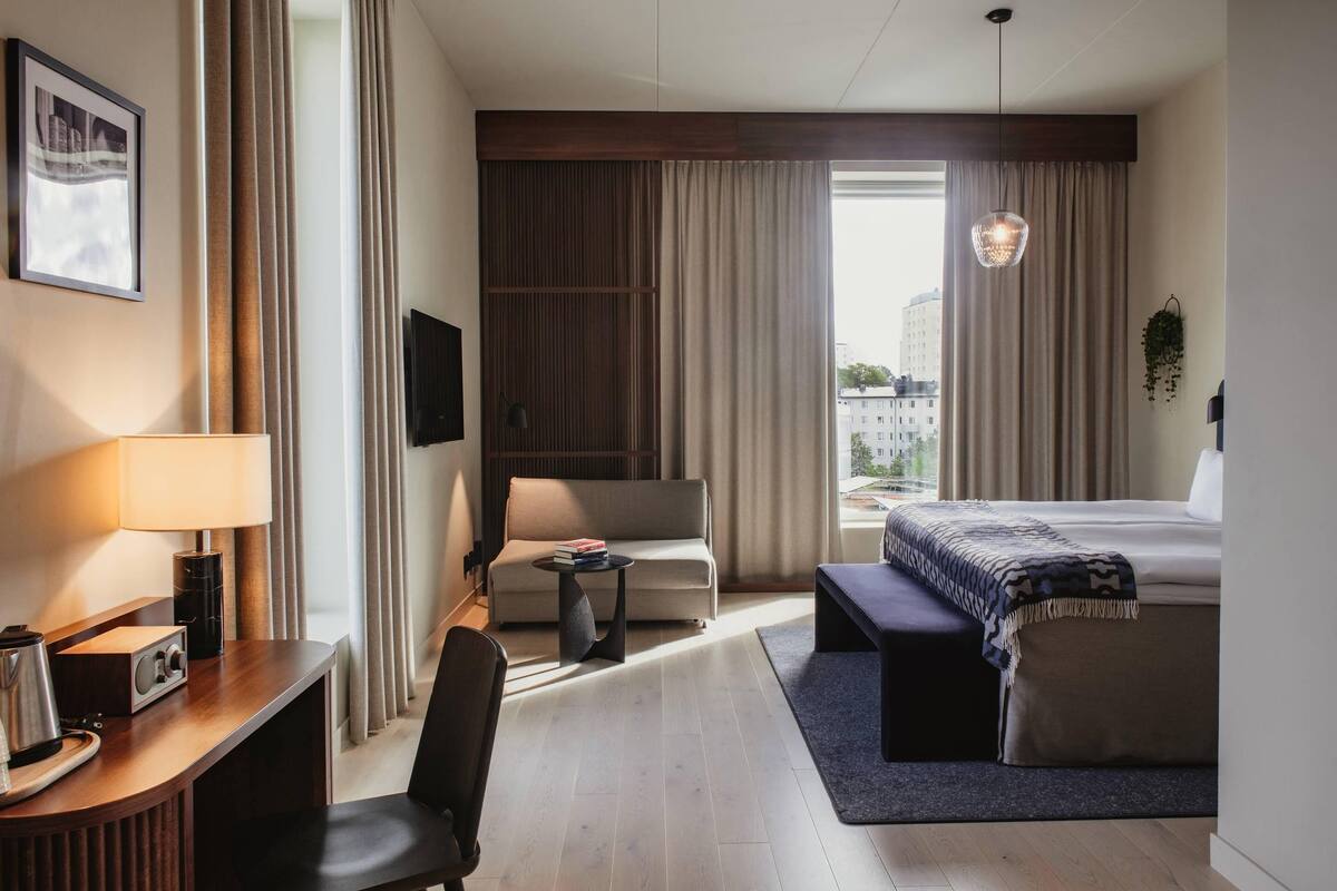 Habitación de hotel con cama, sofá, escritorio con lámpara, silla y una mesita con libros.