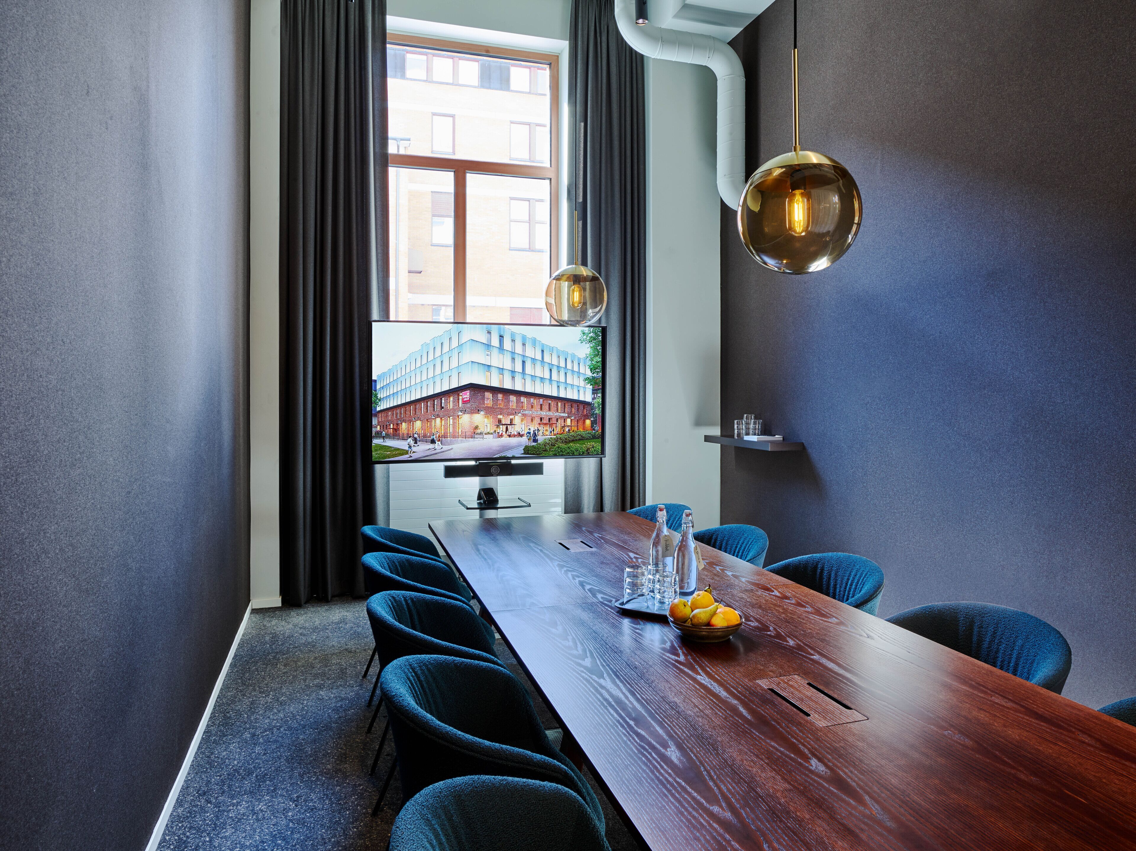 A modern conference room with a long wooden table, blue upholstered chairs, a large flat-screen TV displaying an exterior view, and a pendant light.