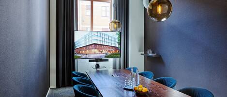 A modern conference room with a long wooden table, blue upholstered chairs, a large flat-screen TV displaying an exterior view, and a pendant light.