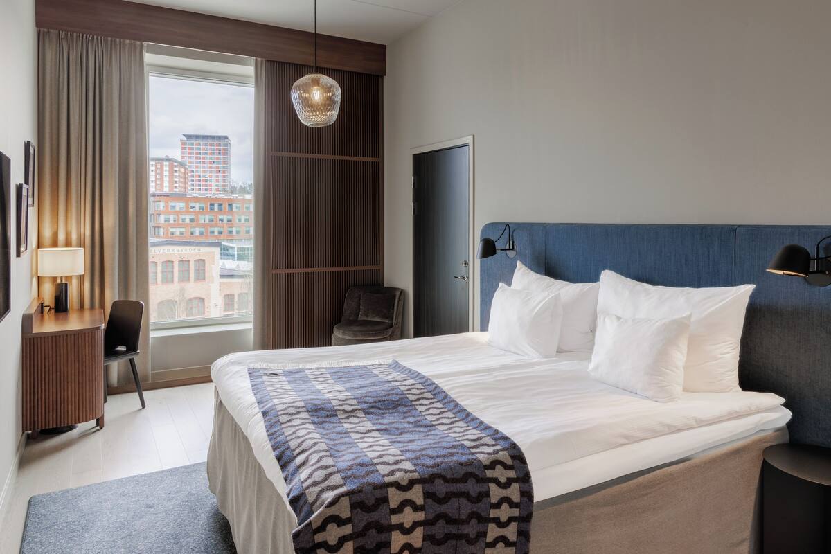 Habitación de hotel con una cama grande, un escritorio con lámpara, una silla, un armario y vistas a edificios a través de una ventana.