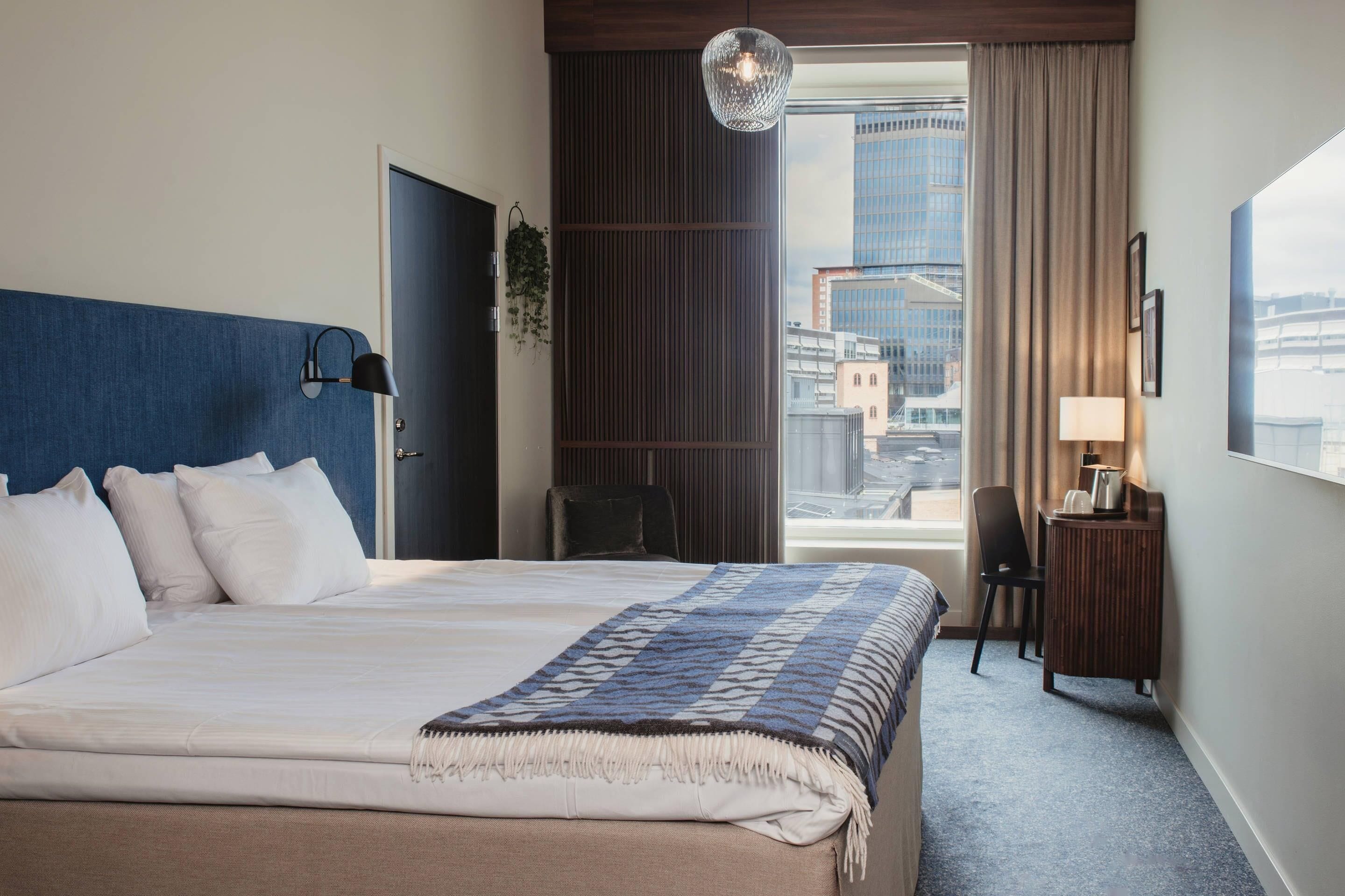 A hotel room with a large bed, a desk, a chair, a window with a view of buildings, and a hanging light fixture.