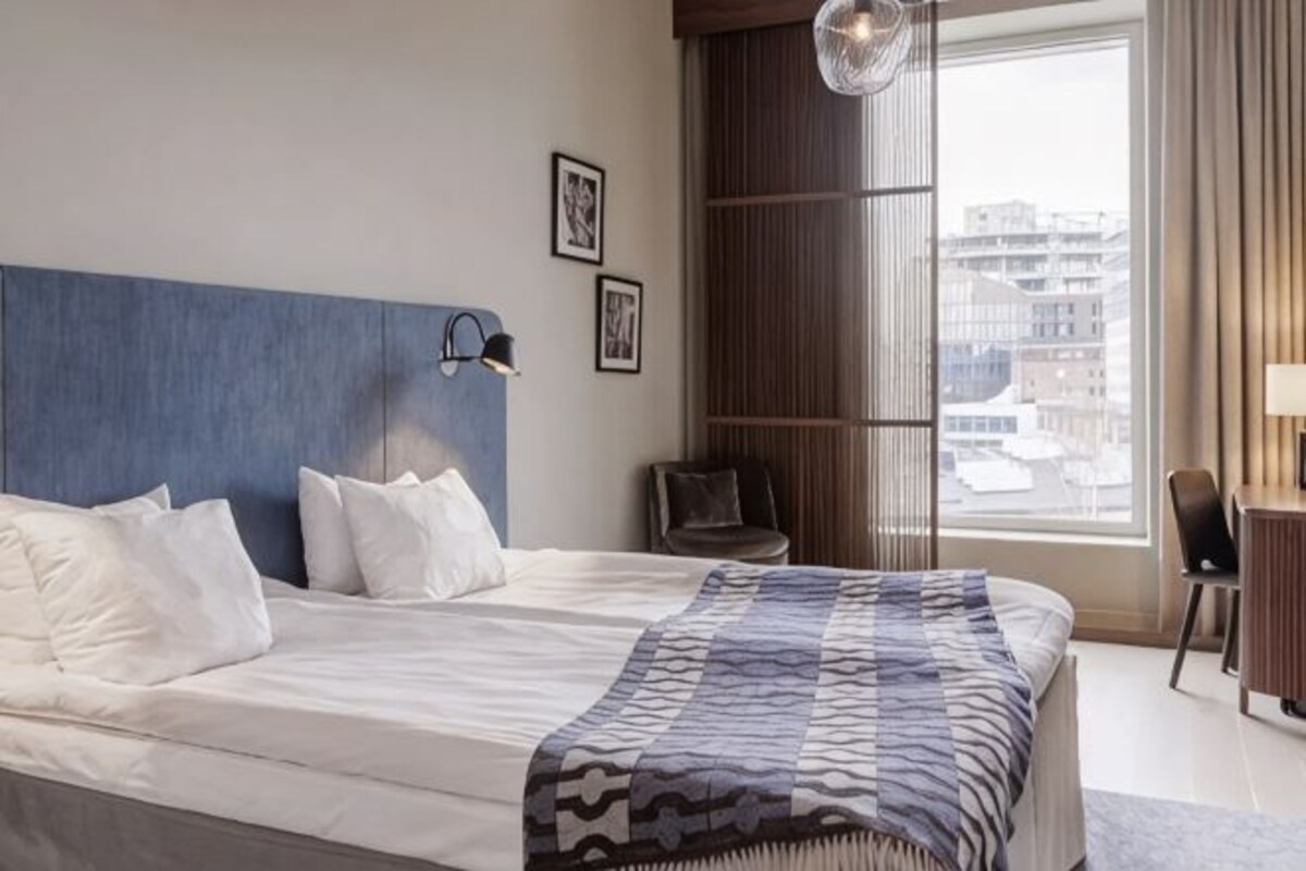 A hotel room with a large bed, a desk, a chair, a window with a view of buildings, and a wall-mounted lamp.