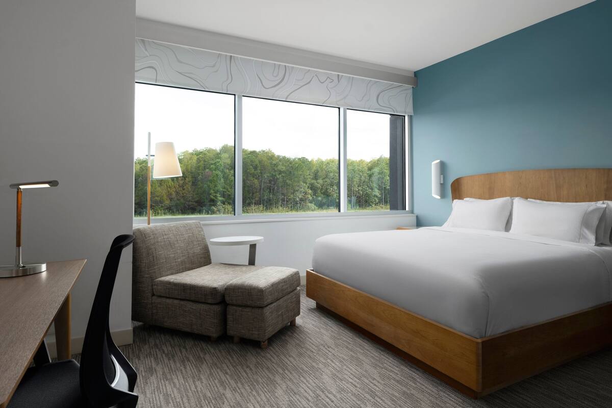 Een moderne hotelkamer met een groot bed, een bureau met lamp, een stoel en uitzicht op het bos door grote ramen.