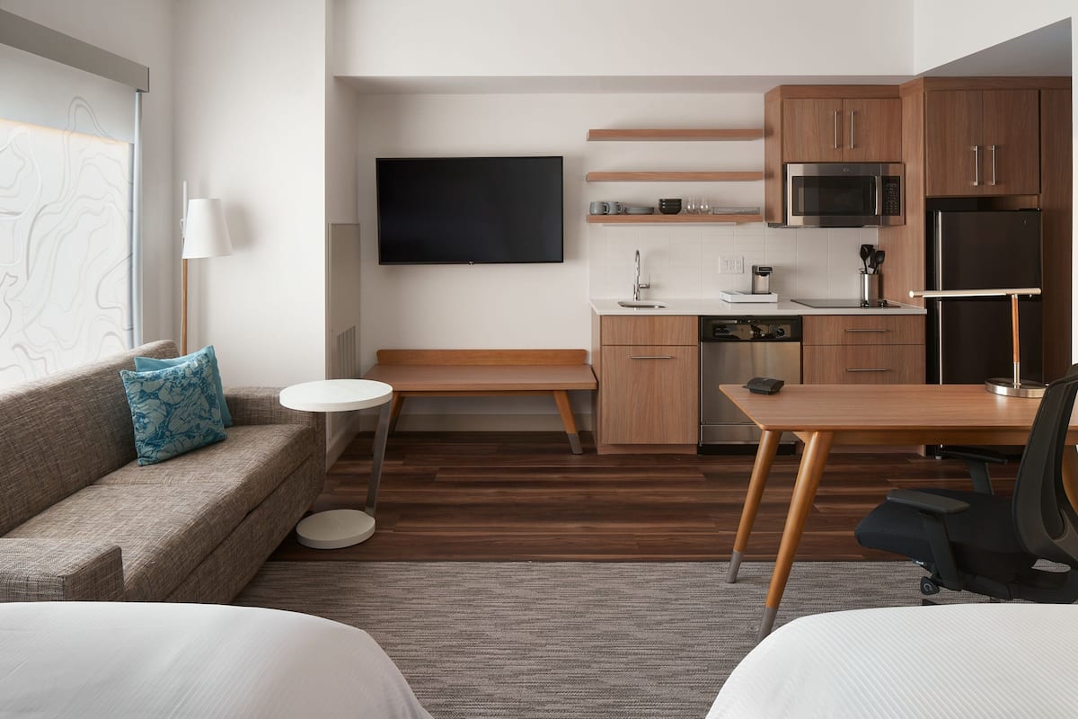 Een moderne hotelkamer met een bank, een klein rond tafeltje, een bureau met stoel, een flatscreen televisie aan de muur, een magnetron, een koelkast en houten kasten.