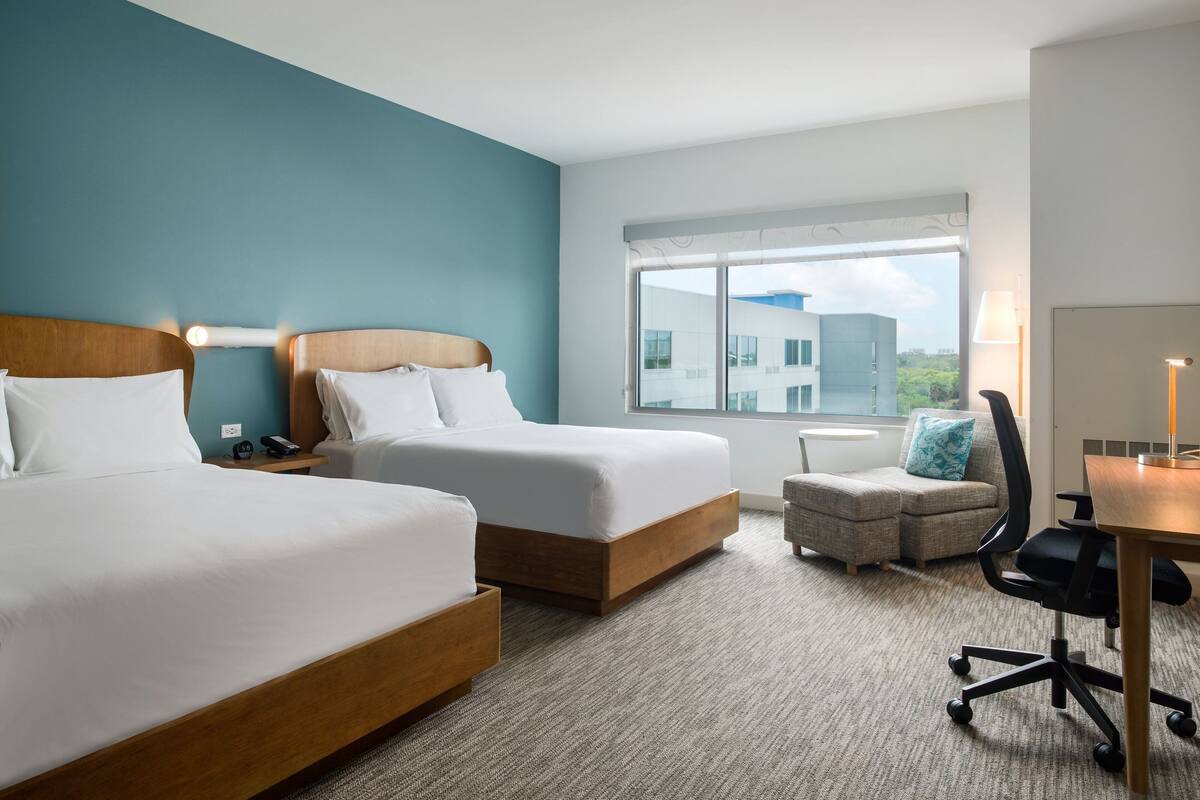 Een hotelkamer met twee bedden, een bureau, een stoel en een raam met uitzicht op gebouwen.