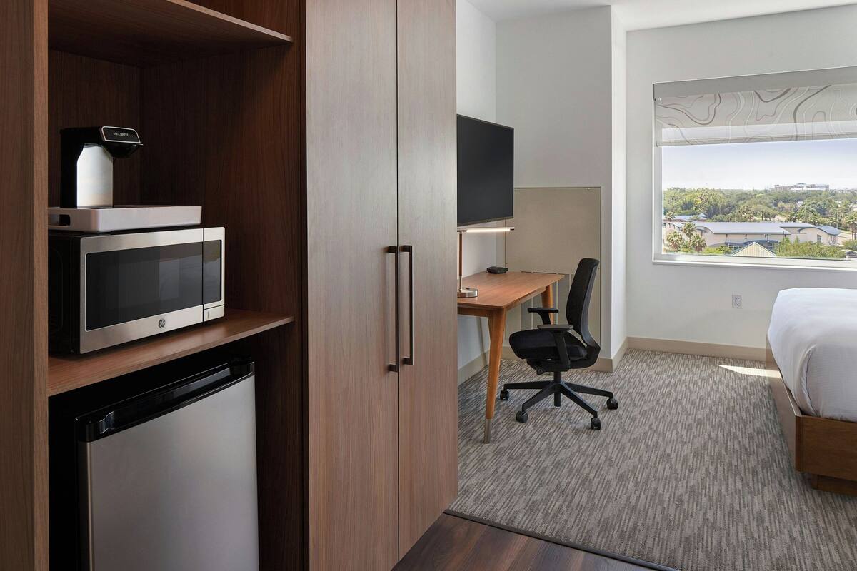 Hotelkamer met kitchenette, bed, bureau met stoel, televisie en raam met uitzicht op gebouwen.