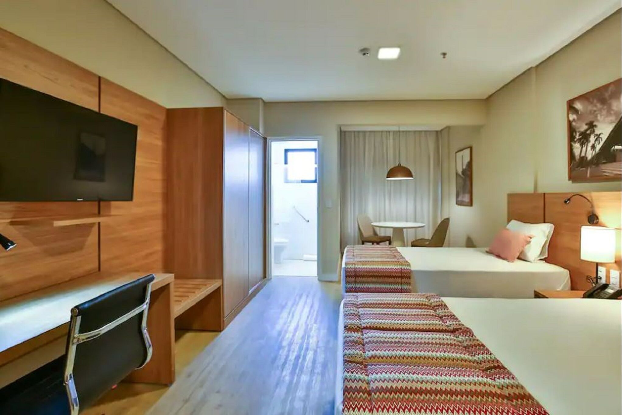 A hotel room with a TV, a desk, a bed, a bedside table, a lamp, and a window with curtains.