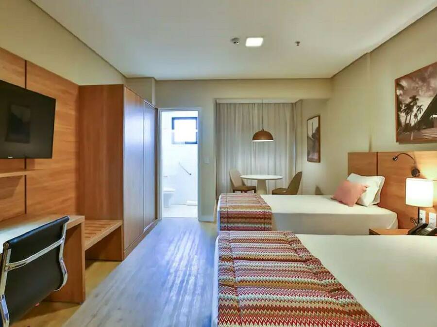 A hotel room with a TV, a desk, a bed, a bedside table, a lamp, and a window with curtains.