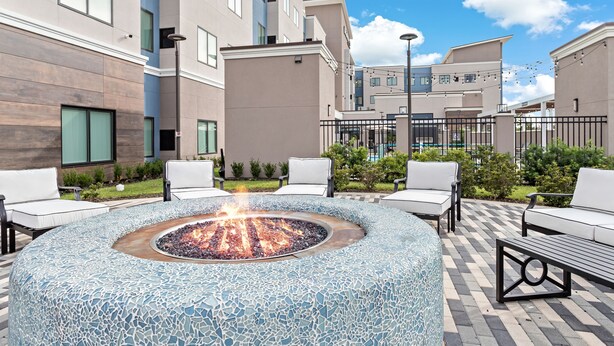 A circular mosaic fire pit surrounded by white outdoor furniture, set against a backdrop of modern buildings and a clear sky. - Residence Inn by Marriott Port Arthur (Port Arthur)