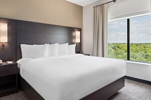 Habitación de hotel con una cama grande, una mesita de noche, una lámpara y una ventana con vistas a zonas verdes.