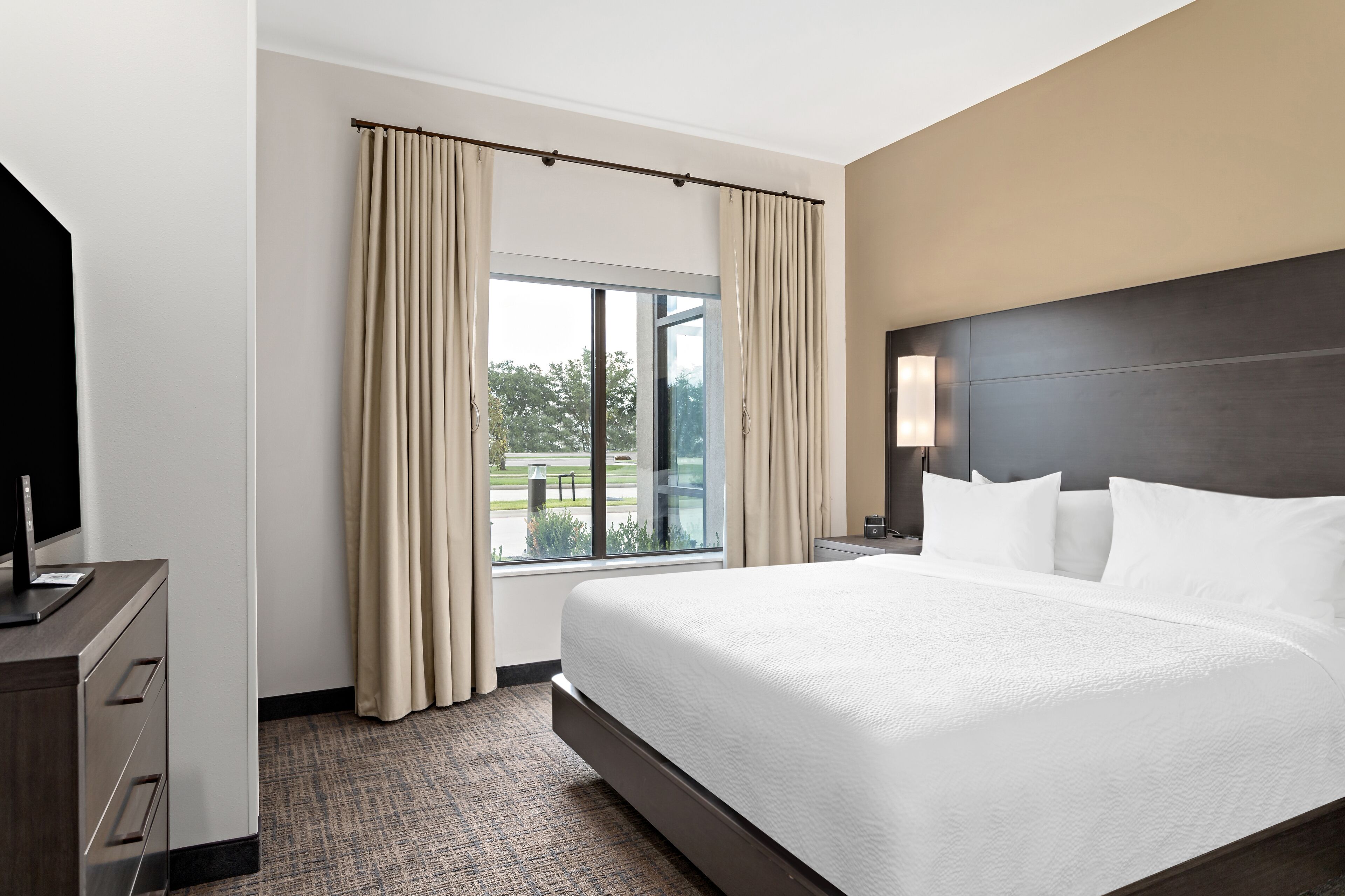 A hotel room with a large bed, a desk with a computer, a window with curtains, and a view of the outdoors.