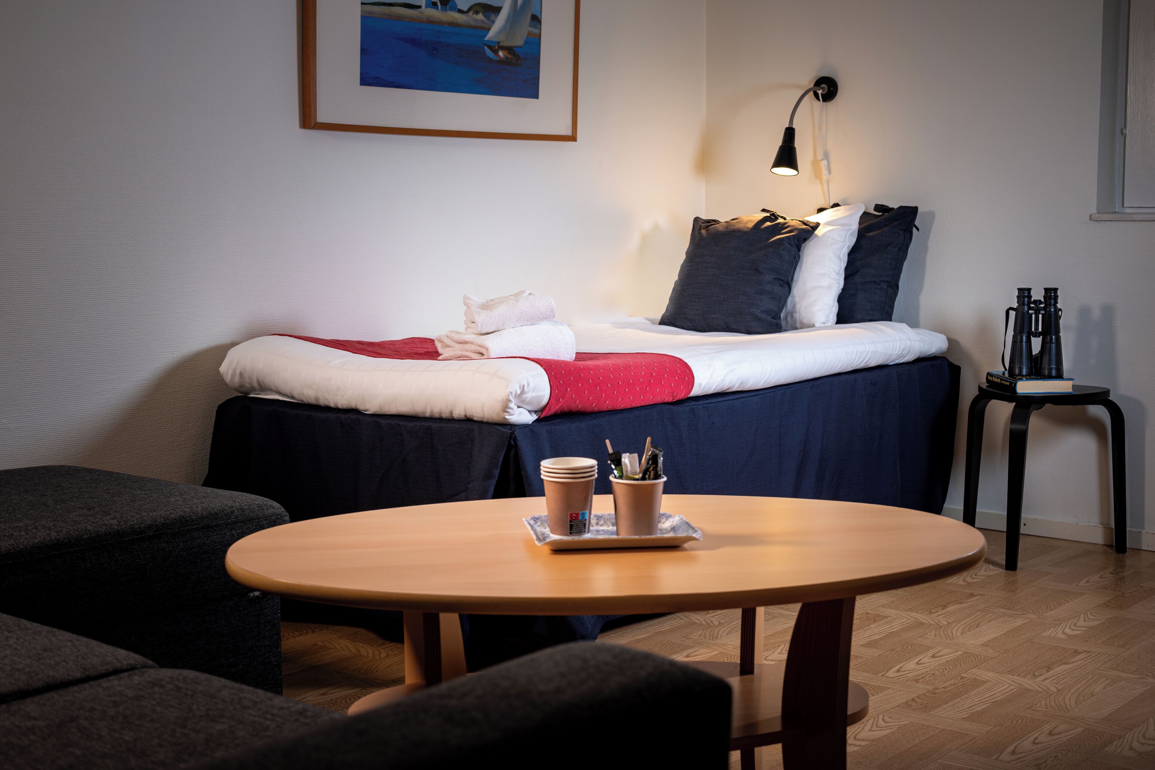 Quarto de hotel bem arrumado, com cama, uma mesinha com um tabuleiro de objetos, uma cadeira e uma mesa de cabeceira com abajur.