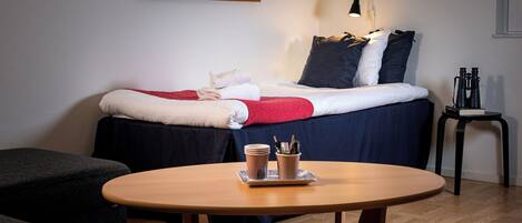 Quarto de hotel bem arrumado, com cama, uma mesinha com um tabuleiro de objetos, uma cadeira e uma mesa de cabeceira com abajur.