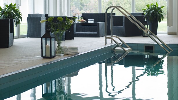 Un área de piscina interior moderna con paredes de vidrio transparente, piso de baldosas y una zona de estar con muebles oscuros y plantas en macetas.