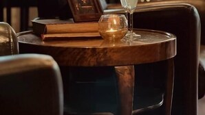 Una mesa con un libro, una vela y un vaso de agua en una habitación poco iluminada, con una planta al fondo.
