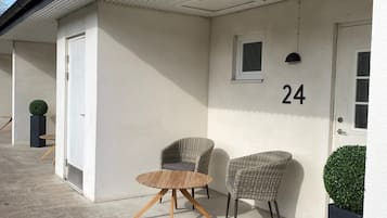En byggnad med ett vågformat metalltak, en vit dörr med nummer 24, två flätade fåtöljer och ett runt träbord.