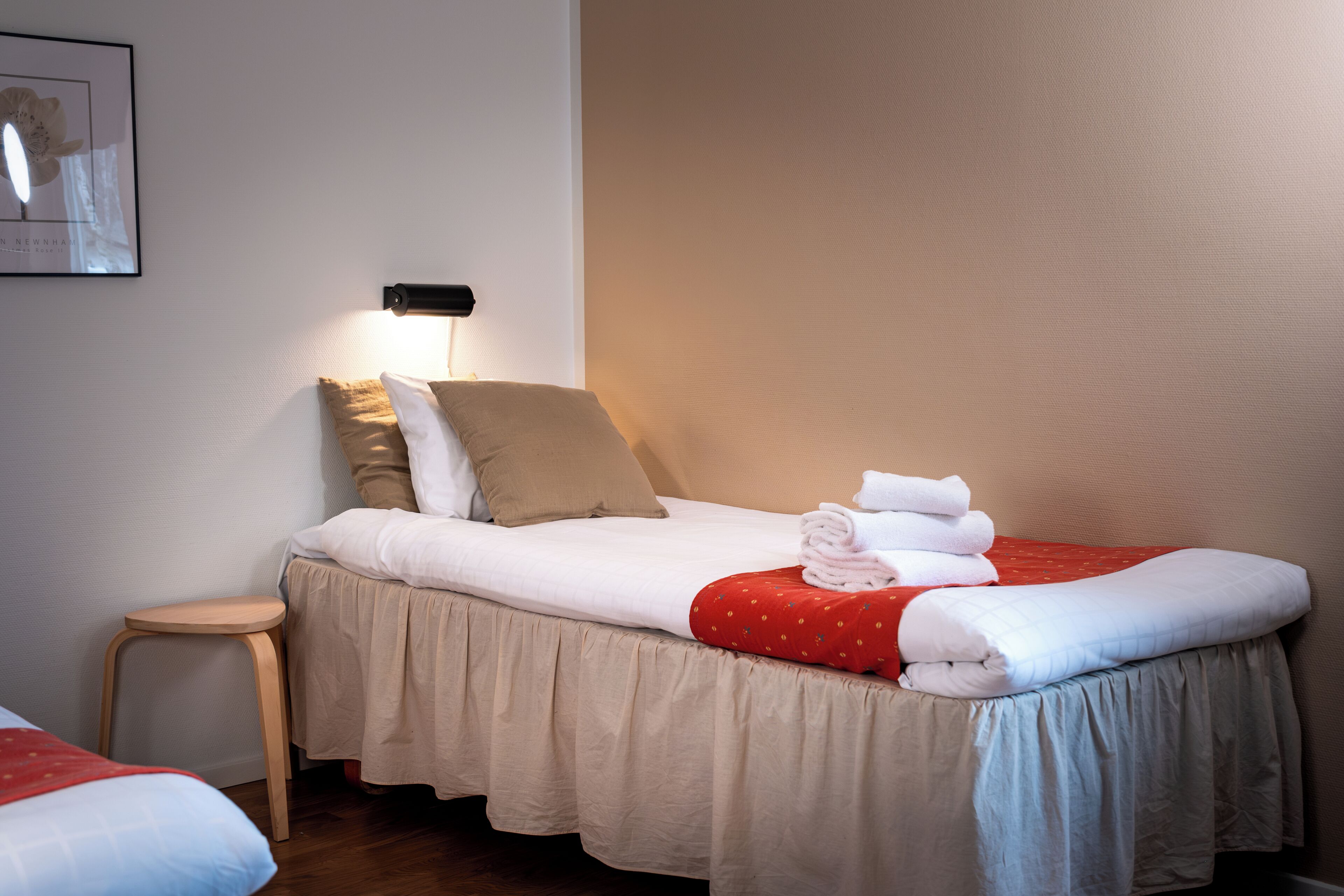 Una cama bien hecha con una colcha roja y blanca, una falda para cama color beige y un taburete cerca.