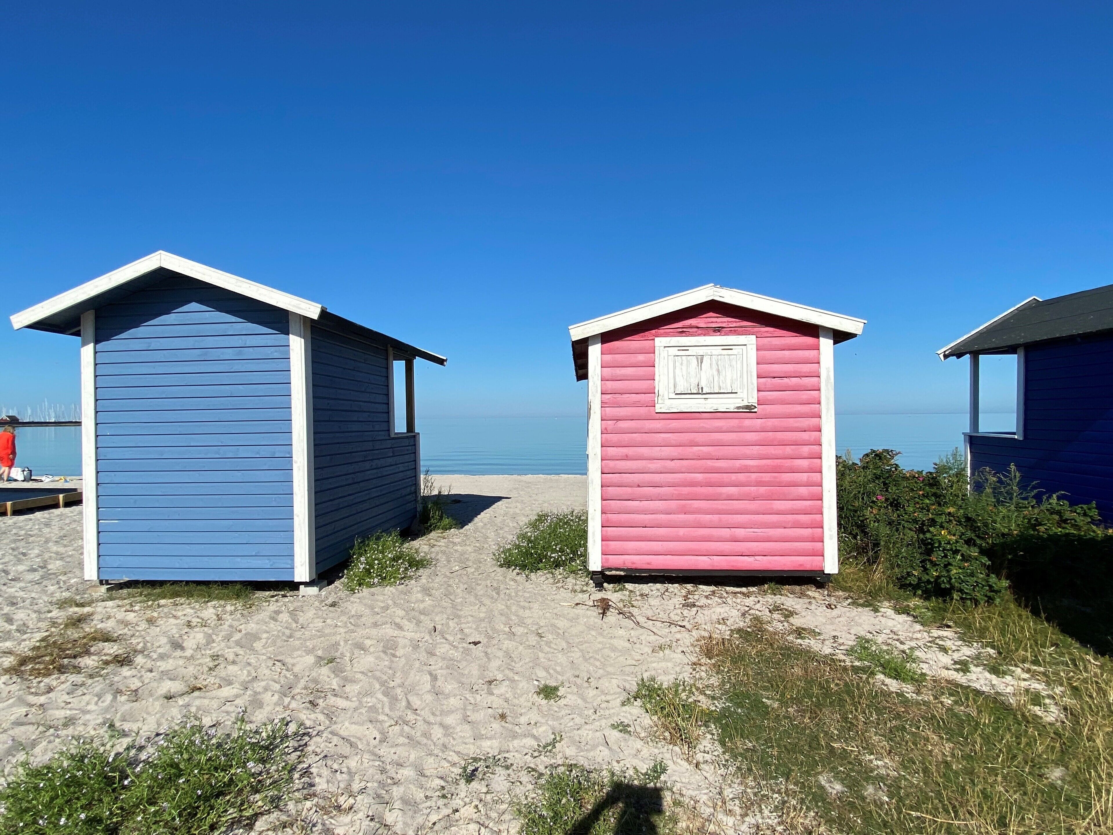 Two beach huts, one blue and one pink, with a clear view of the sea in the background.