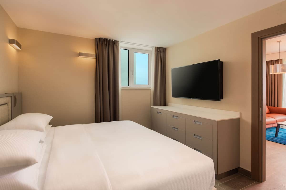 A hotel room with a bed, a TV mounted on the wall, a window with curtains, and a door leading to a seating area.