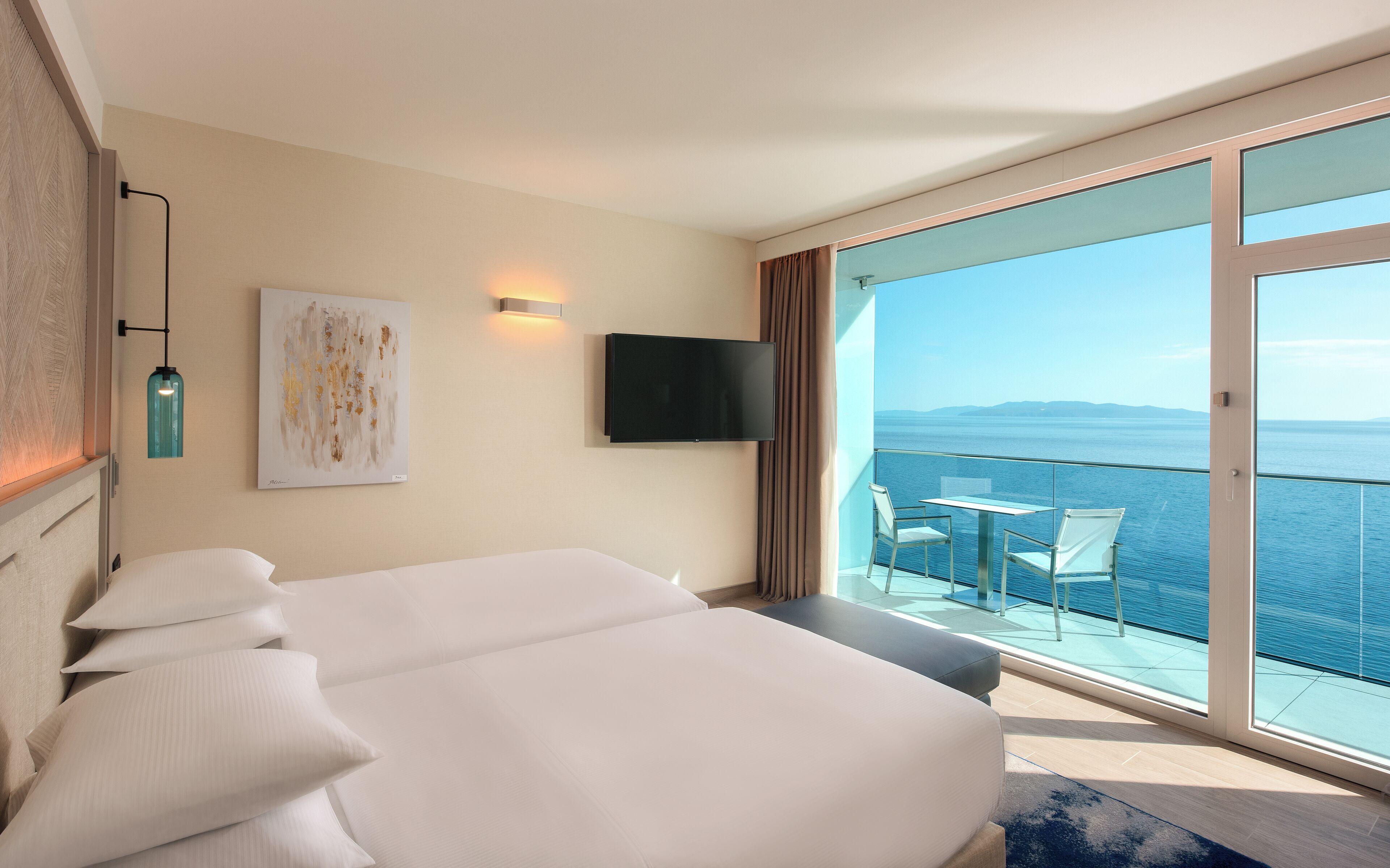 A hotel room with a bed, a TV, a painting, and a balcony with a view of the sea.
